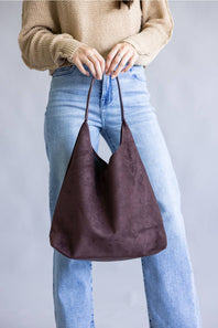 A person wearing a beige knit sweater and light blue jeans holds a large, slouchy hobo shoulder bag in dark brown suede against a plain gray studio background.