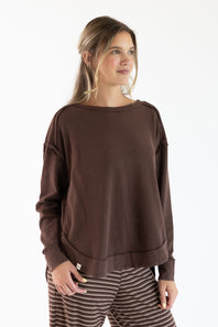 A woman wearing a dark brown long-sleeve crewneck sweatshirt and brown-and-white striped pants, photographed against a clean white studio background.