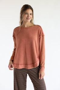 A woman modeling a washed, dusty terracotta long-sleeve top and brown-and-white striped pants against a plain white studio background.