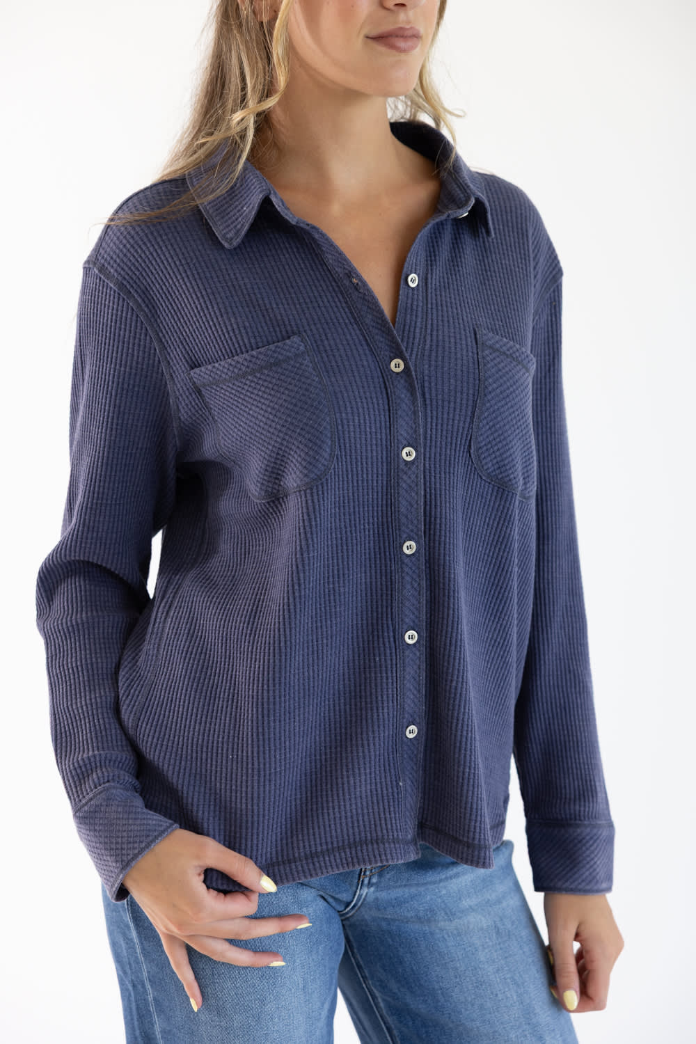 A woman wearing a navy blue ribbed long-sleeve button-up shirt with chest pockets, paired with blue jeans, against a plain white background.