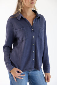 A woman wearing a navy blue ribbed long-sleeve button-up shirt with chest pockets, paired with blue jeans, against a plain white background.