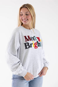A person wearing a light heather gray crewneck sweatshirt with the words 'Merry & Bright' printed across the chest in multicolored textured letters.