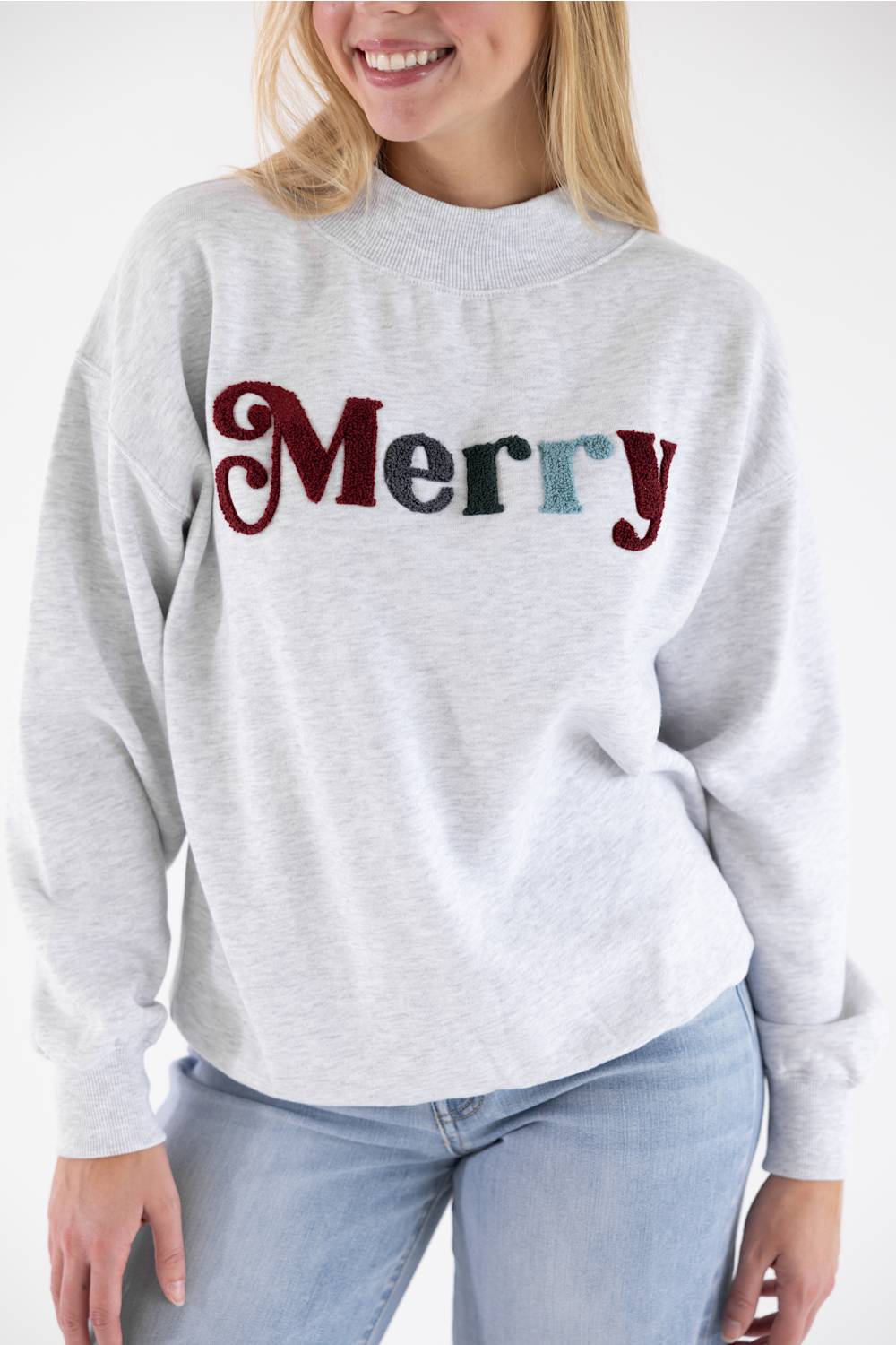 A woman wearing a light heather gray crewneck sweatshirt with 'Merry' embroidered across the chest in red, dark green, and blue sequins, paired with light blue jeans.