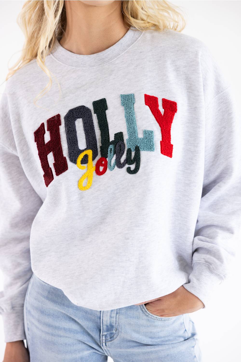 A woman wearing a light heather gray sweatshirt with a colorful 'HOLLY Jolly' graphic on the front, paired with light blue jeans.