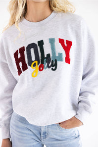 A woman wearing a light heather gray sweatshirt with a colorful 'HOLLY Jolly' graphic on the front, paired with light blue jeans.
