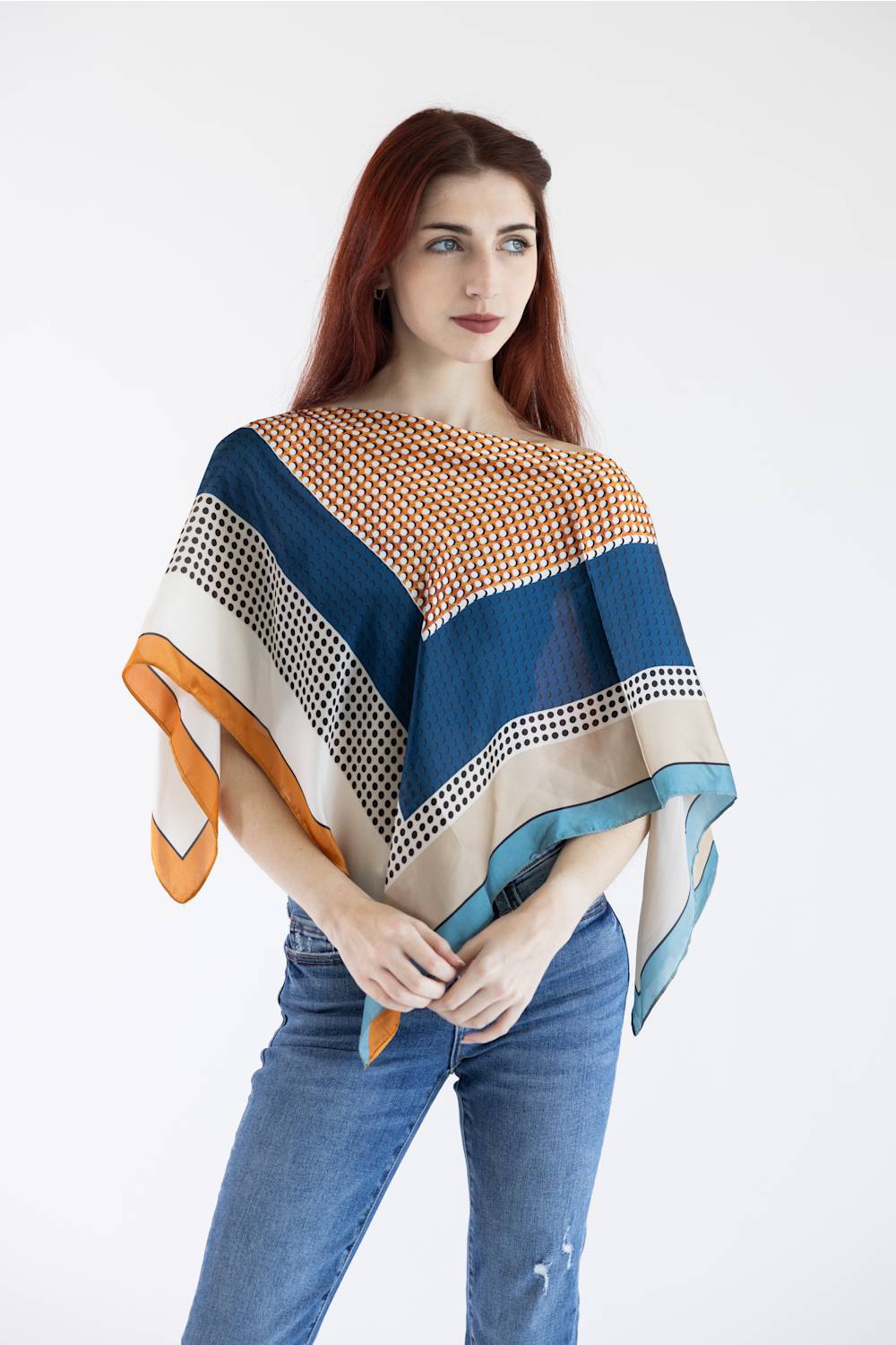A woman with red hair wearing a multi-colored striped and patterned poncho top with blue jeans, standing against a plain white background.