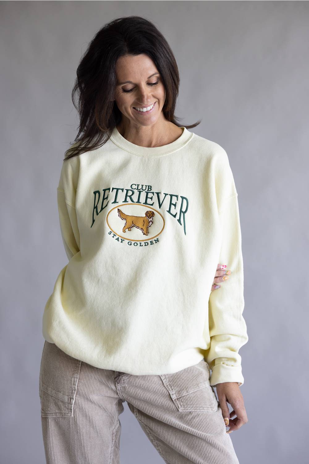 A woman wearing a cream-colored crewneck sweatshirt with a 'Club Retriever Stay Golden' graphic featuring a golden retriever dog, paired with beige corduroy pants, against a neutral gray studio background.