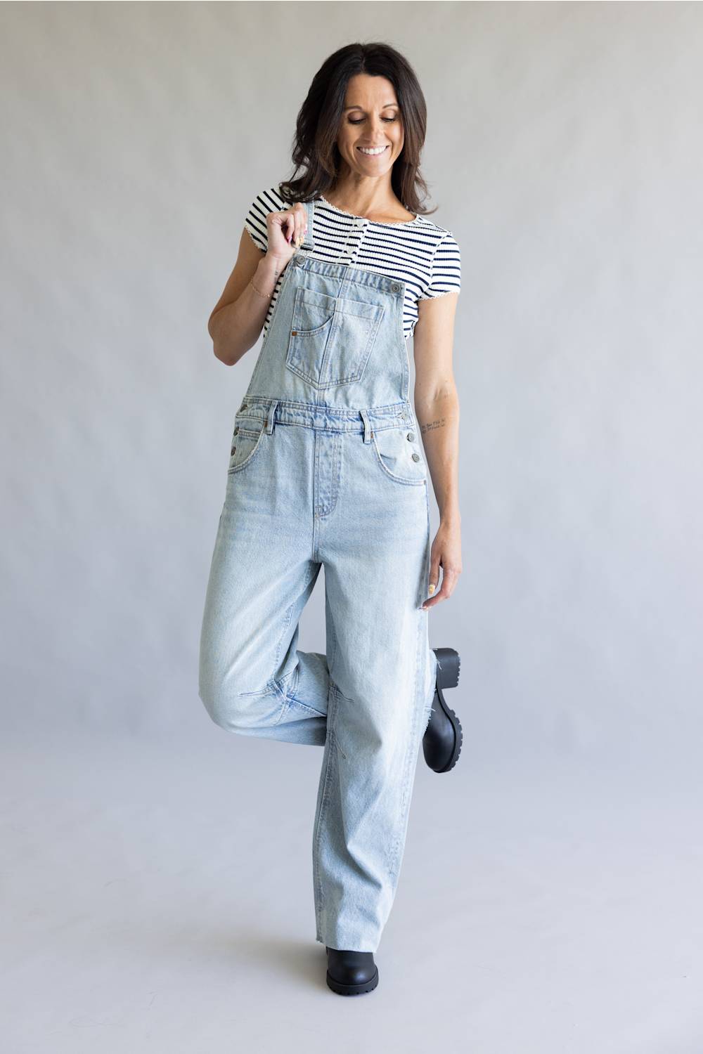 A woman modeling light-wash denim overalls over a black and white striped t-shirt, standing against a neutral light gray studio background.