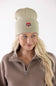 A woman with blonde hair smiling while wearing a faded olive green Carhartt watch cap beanie featuring a small red heart patch on the front fold.