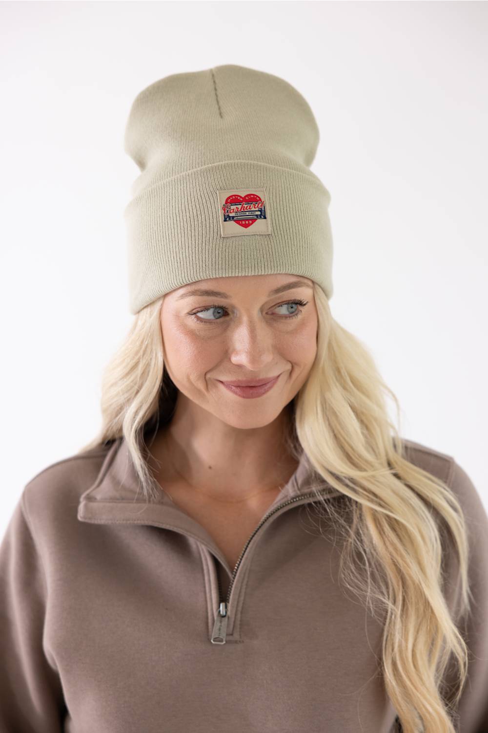 A woman with blonde hair smiling while wearing a faded olive green Carhartt watch cap beanie featuring a small red heart patch on the front fold.