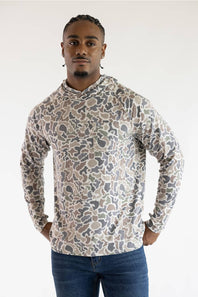 A man wearing a long-sleeve camouflage hoodie with a beige, gray, and brown camo pattern, paired with blue jeans, standing against a plain white studio background.