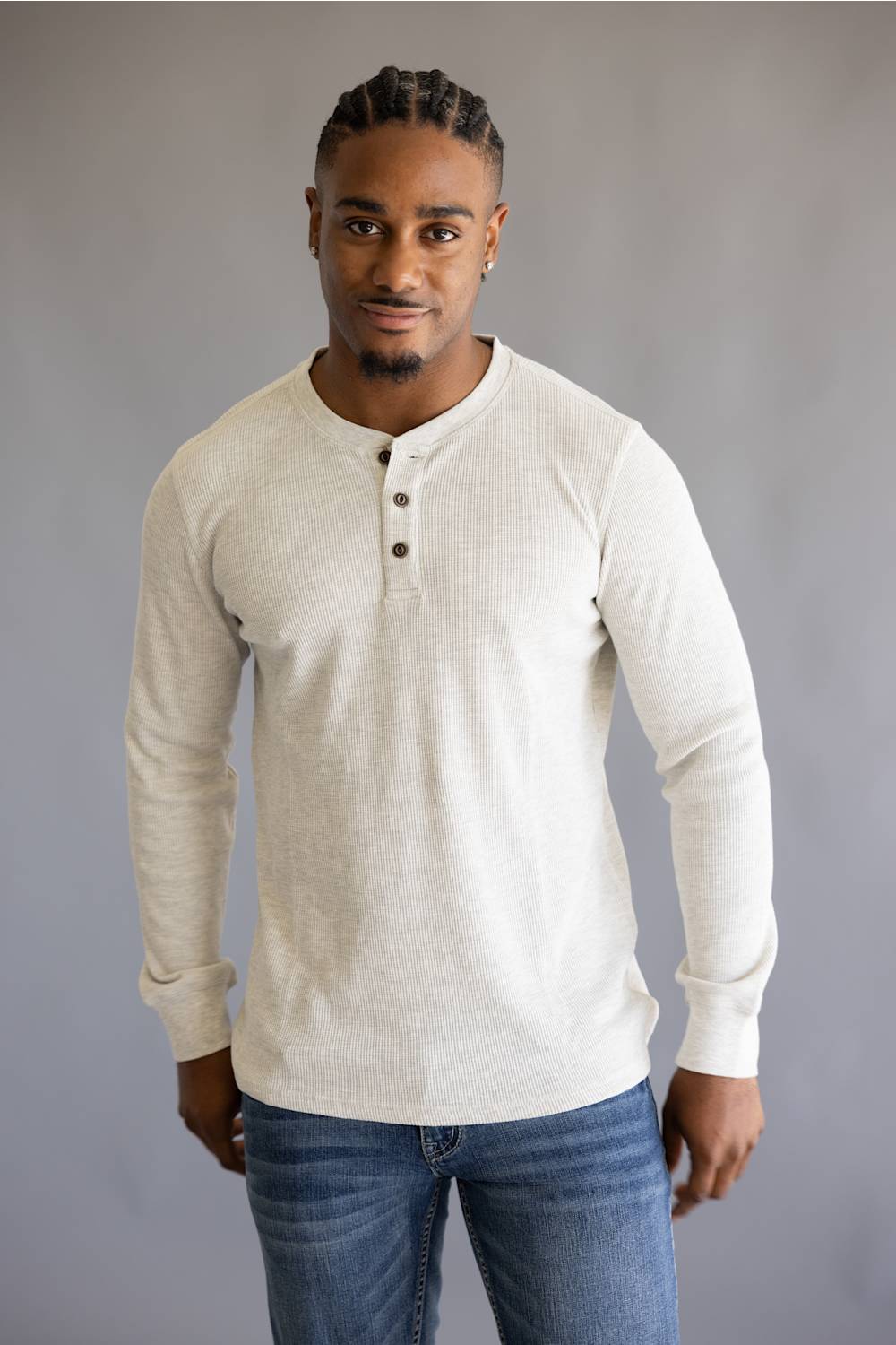 A man wearing a light beige long-sleeve henley shirt with three buttons on the chest, paired with blue jeans, standing against a neutral gray background.