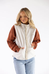 A woman wearing a two-tone fleece jacket with a cream body and brown sleeves, paired with light blue jeans, against a plain white background.