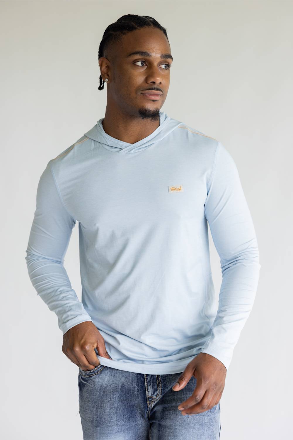 A man wearing a light blue long-sleeve hooded shirt with a small gold chest logo, paired with blue jeans, photographed against a plain light gray background.