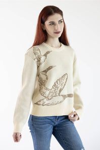 A woman with red hair wearing a cream-colored knit sweater with a brown graphic of flying ducks, paired with blue jeans, against a plain white background.