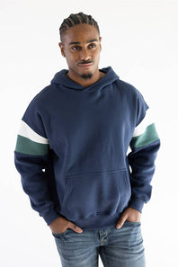 A man wearing a navy blue hoodie with green and white color-blocked sleeves, paired with blue jeans, standing against a plain white background.