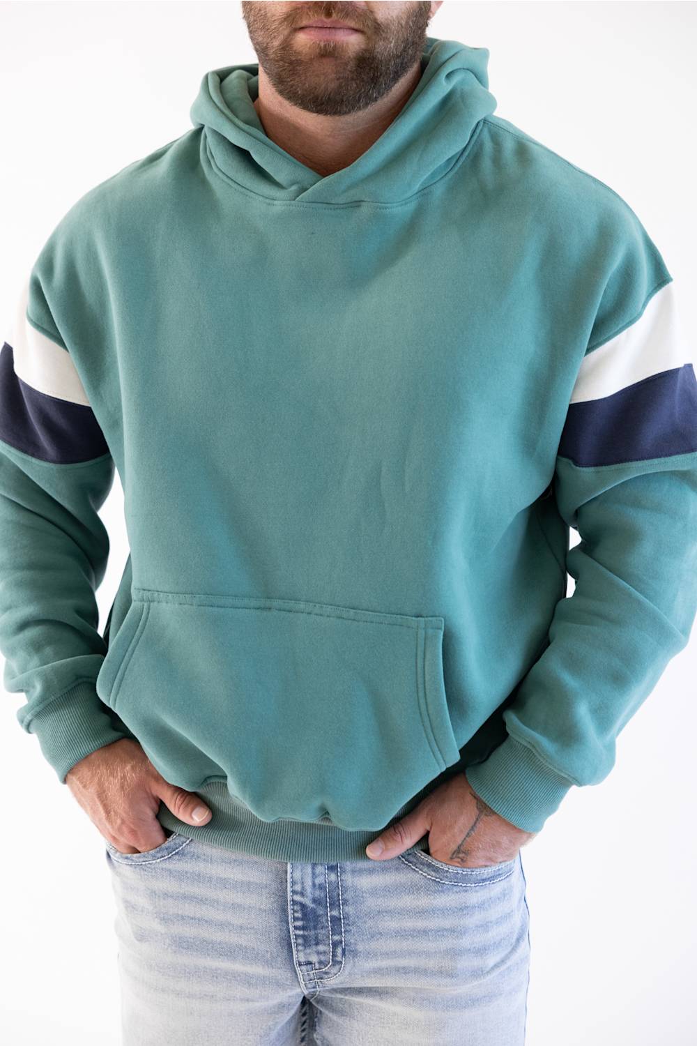 A man wearing a teal green hooded sweatshirt with white and navy blue stripes on the sleeves, paired with light-wash blue jeans, against a plain white background.