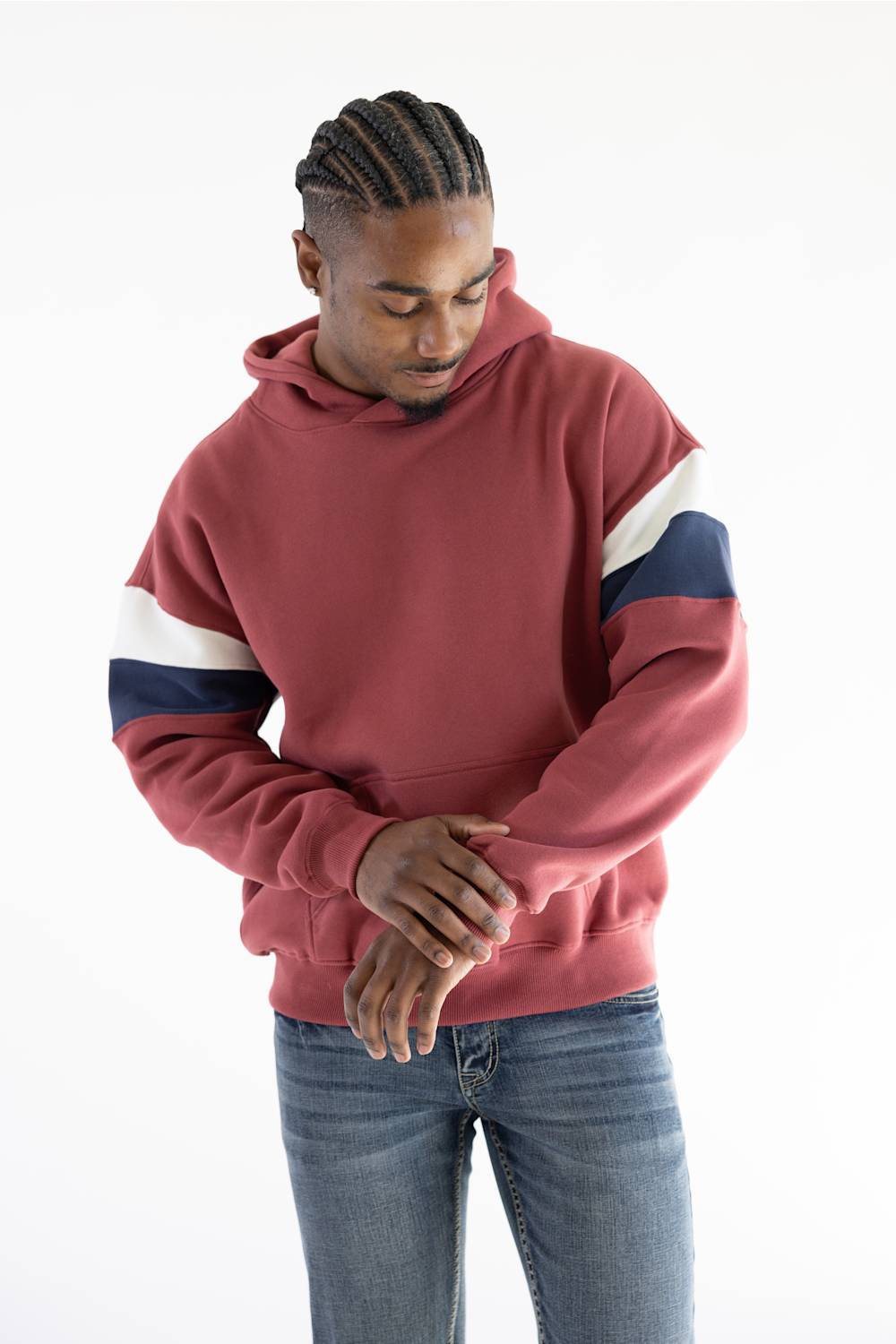 A man wearing a dusty rose red hoodie with white and navy blue block stripes on the sleeves, paired with blue jeans, standing against a plain white background.