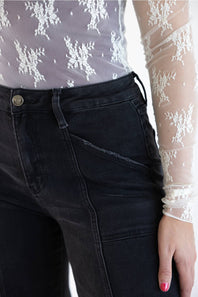 Close-up of a person wearing black denim jeans and a sheer white lace long-sleeve top, with the hand and lower torso visible against a plain light background.