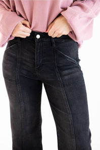 Close-up of a person wearing black denim jeans and a pink ribbed long-sleeve top, with hands pulling the hem up slightly at the waistband against a plain white background.