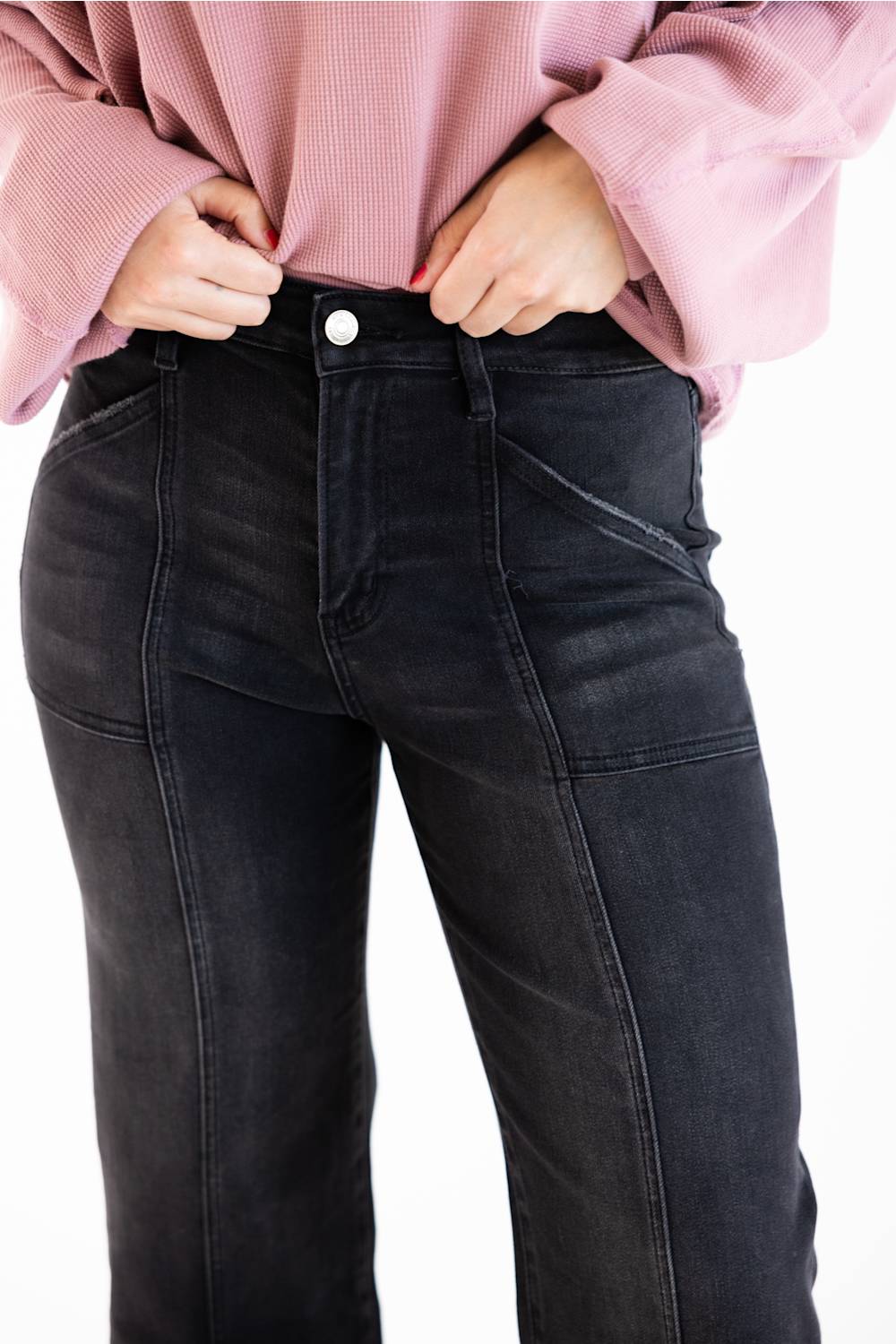 Close-up of a person wearing black denim jeans and a pink ribbed long-sleeve top, with hands pulling the hem up slightly at the waistband against a plain white background.