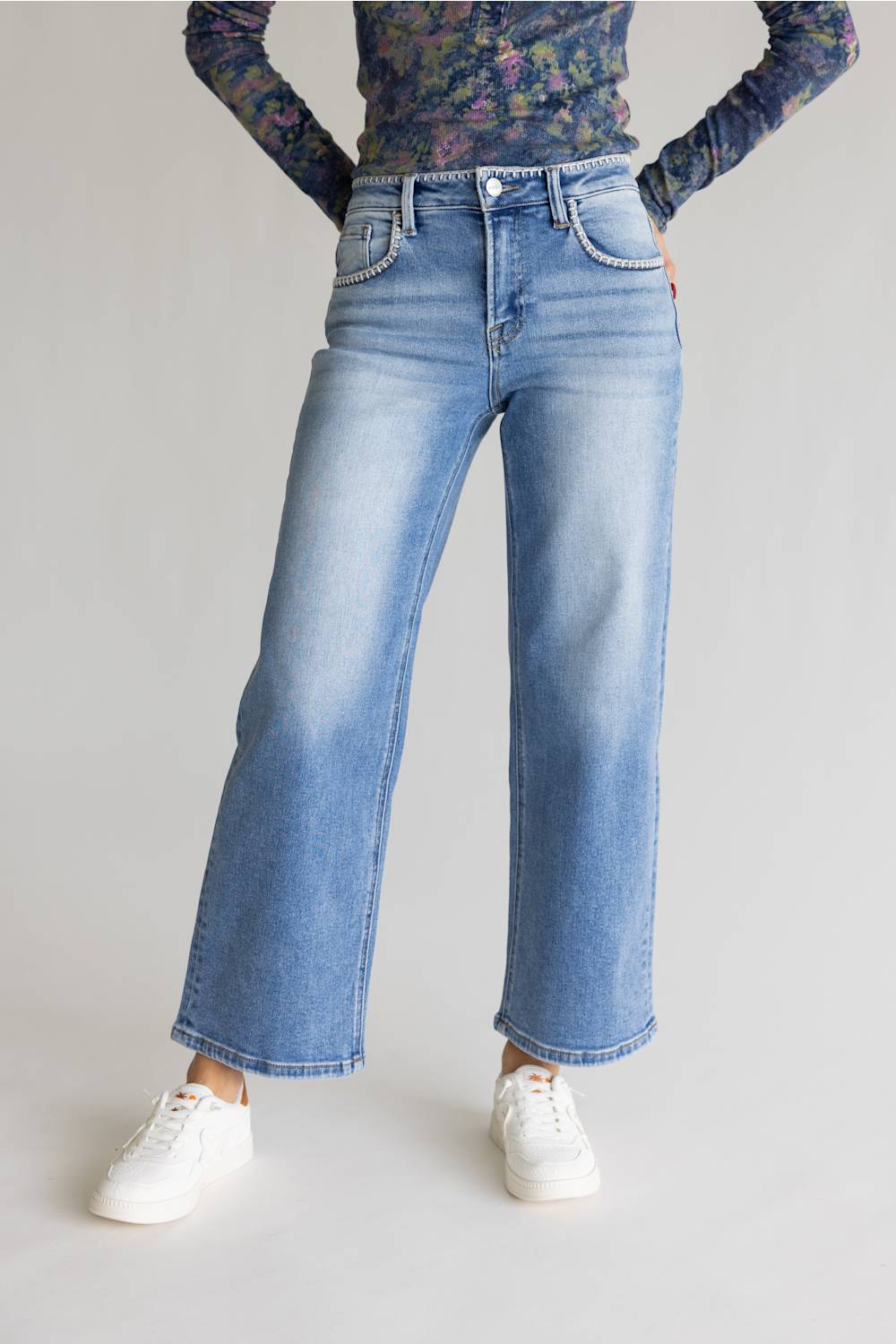 A person wearing light-wash blue wide-leg denim jeans, a dark floral long-sleeve top, and white sneakers, standing against a plain light gray studio background.