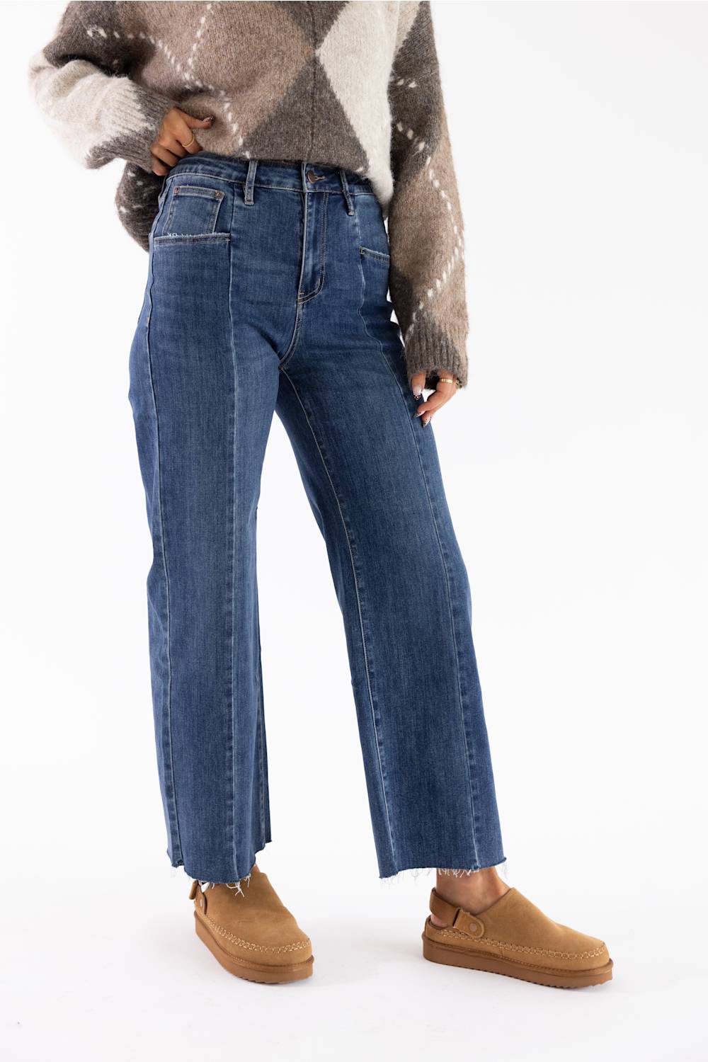 A person wearing blue wide-leg cropped jeans, a brown and beige argyle sweater, and tan slip-on shoes against a plain white studio background.