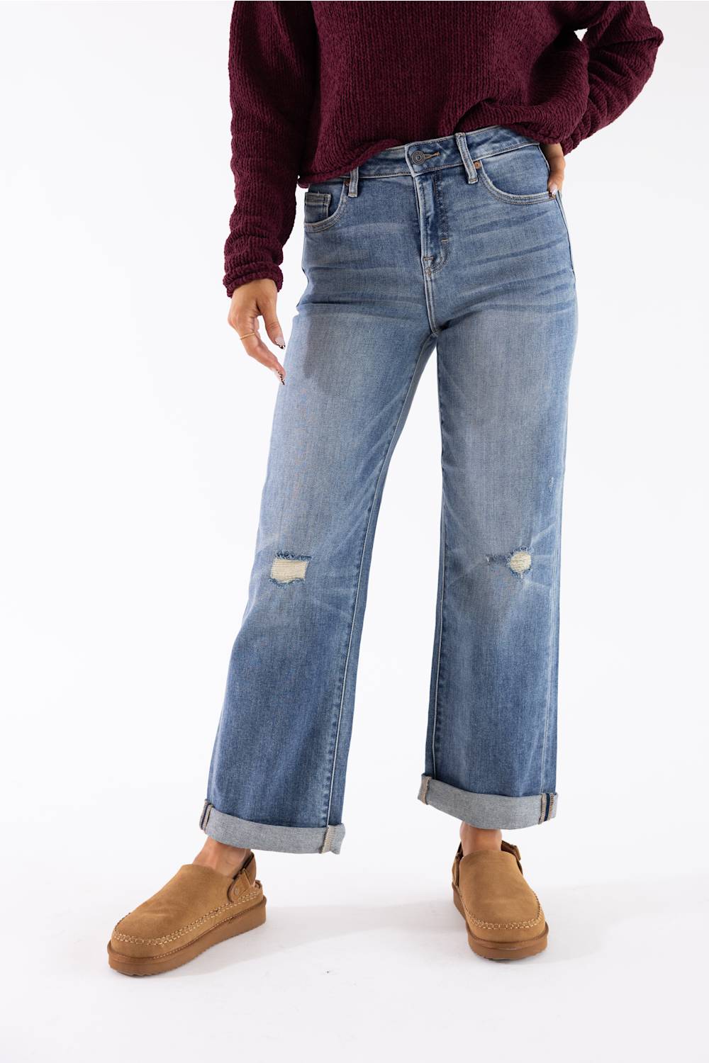 A person wearing light blue wide-leg distressed jeans with cuffed hems, paired with a dark burgundy sweater and tan slip-on shoes, shown against a plain white studio background.