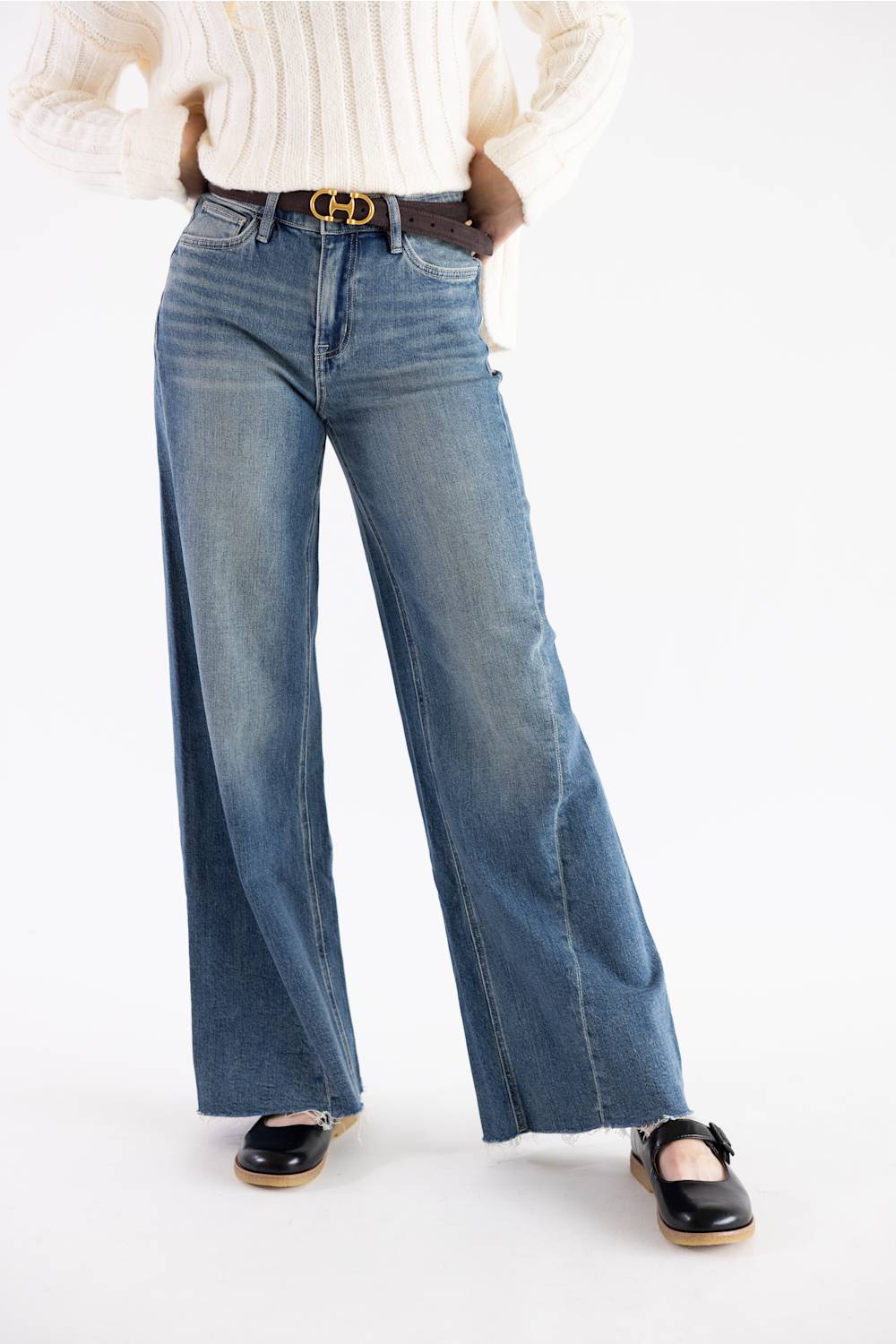 A person wearing blue wide-leg denim jeans with a brown leather belt featuring a gold-tone double-H buckle, styled with a cream ribbed sweater and black Mary Jane shoes against a plain white studio background.