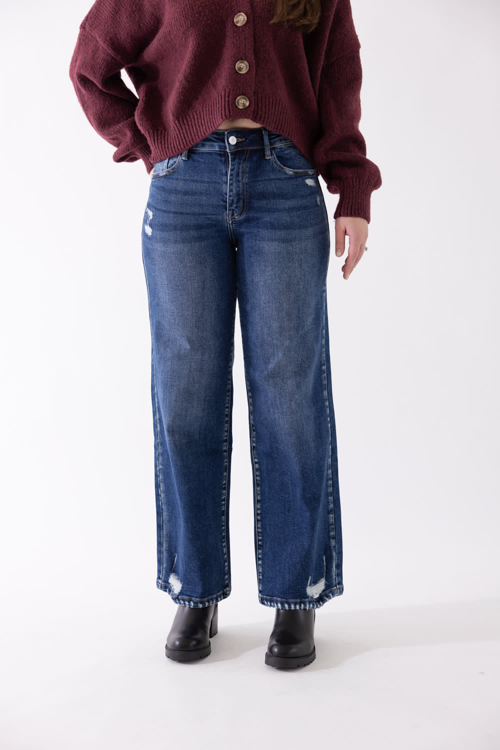 Side view of a model wearing KanCan dark wash, high-rise wide-leg jeans with subtle distressing near the knee and hem, styled with black boots and a burgundy sweater against a plain backdrop.