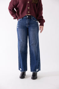 Side view of a model wearing KanCan dark wash, high-rise wide-leg jeans with subtle distressing near the knee and hem, styled with black boots and a burgundy sweater against a plain backdrop.