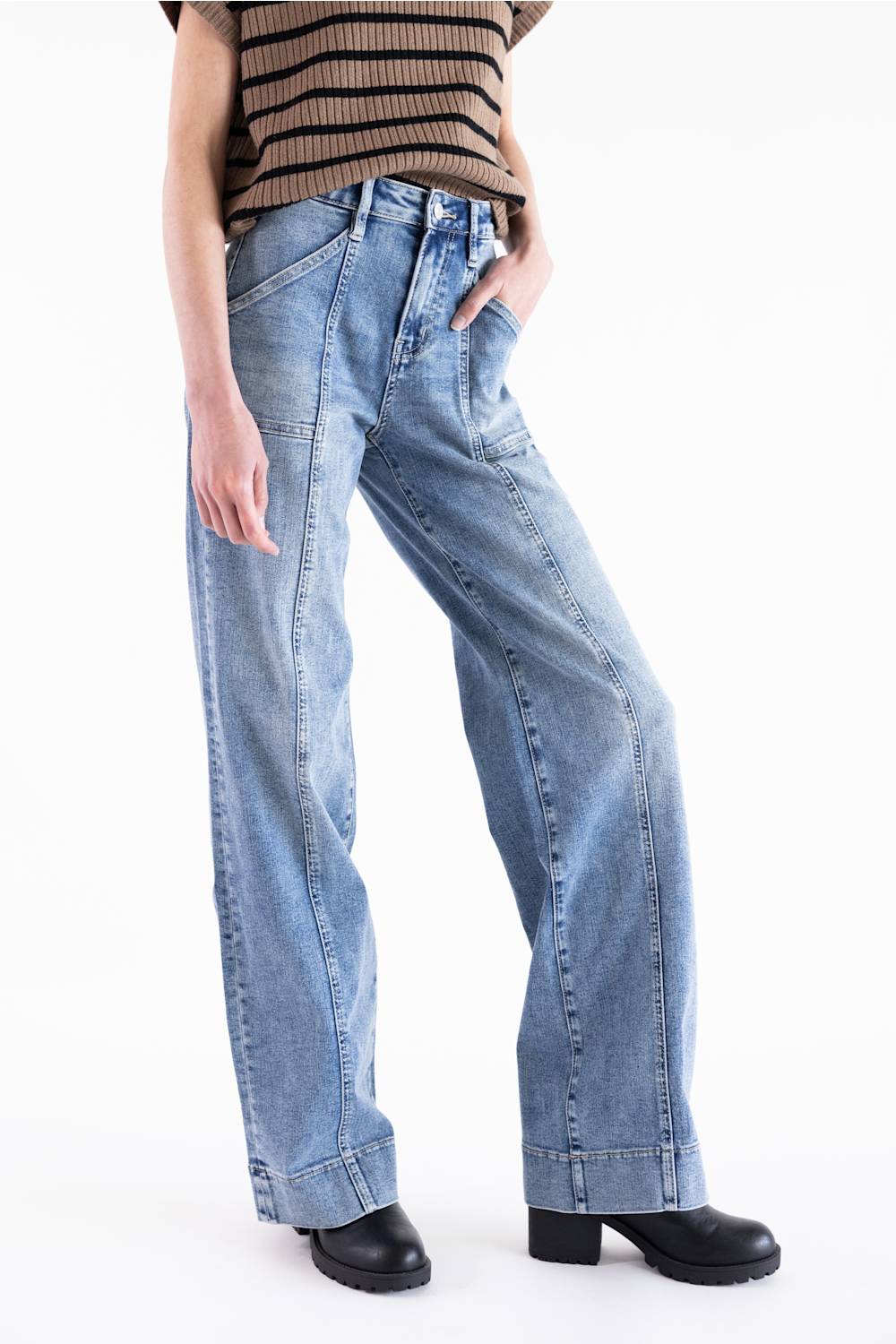 Full-length view of the Mica High Rise Wide Trouser Jeans in light wash, featuring a high-rise waist, distinct front vertical seams, utility pockets, and a wide-leg silhouette. The model is wearing a short-sleeve striped mock neck sweater and black boots against a neutral white background.