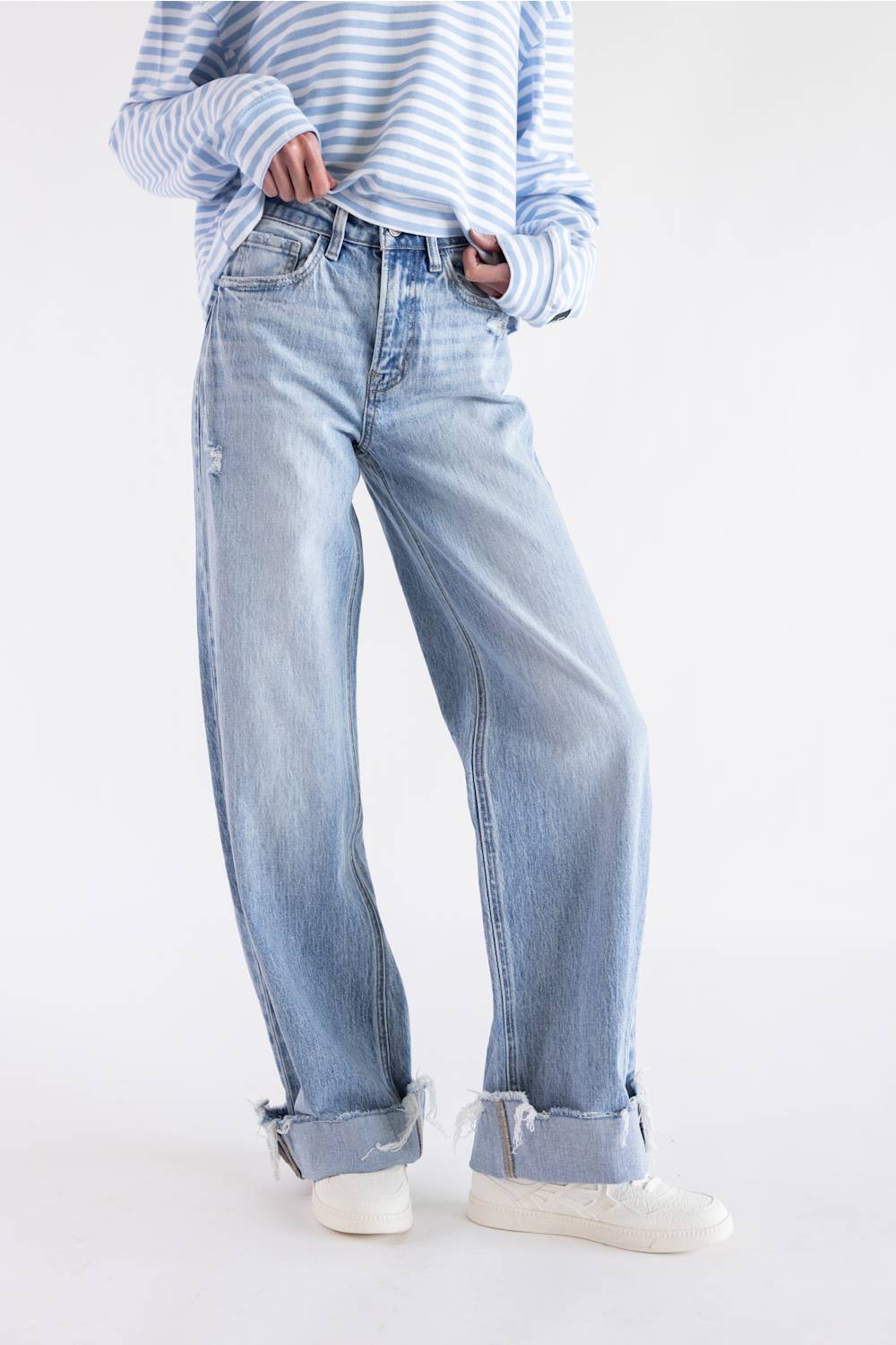A person wearing light-wash blue wide-leg jeans with rolled cuffs, paired with a blue-and-white striped long-sleeve top and white sneakers, photographed against a clean white studio background.