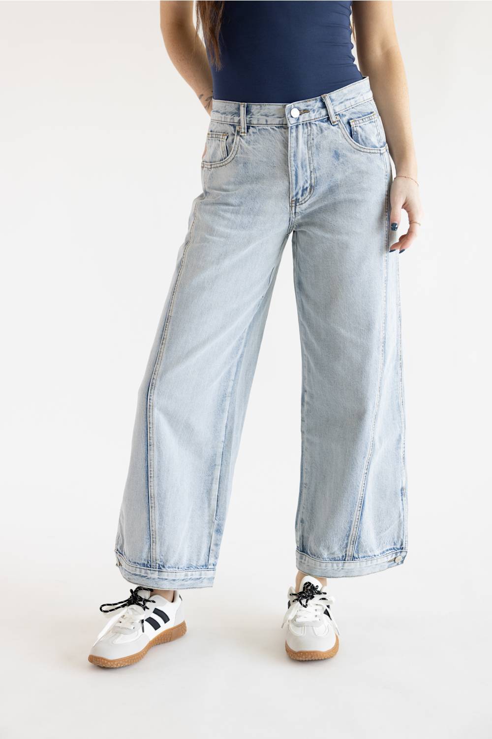 A model posed from the waist down in light-wash wide-leg jeans featuring prominent vertical side seam stitching details, styled with a dark fitted top and white sneakers with brown and black accents on a solid white background.