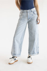 A model posed from the waist down in light-wash wide-leg jeans featuring prominent vertical side seam stitching details, styled with a dark fitted top and white sneakers with brown and black accents on a solid white background.