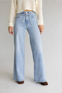 A model wearing light-wash wide-leg denim jeans with a drawstring waist, paired with a cream knit sweater and brown shoes, against a light gray studio background.