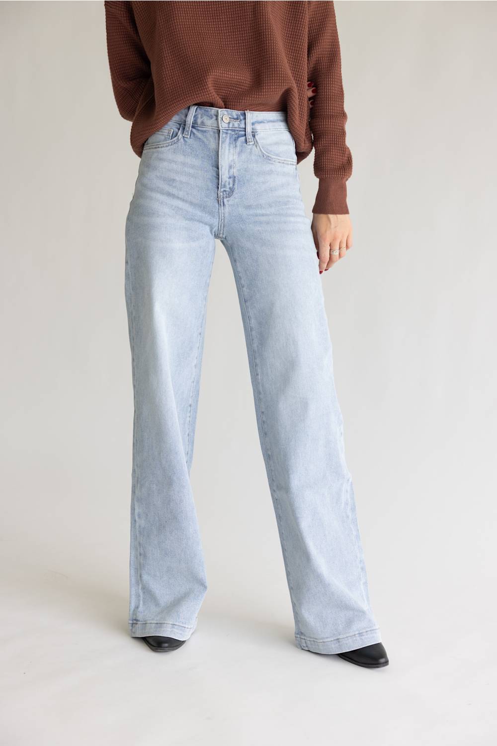 A model wearing light-wash wide-leg denim jeans and a brown knit sweater, standing against a neutral light gray studio background.