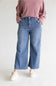 A person wearing blue wide-leg cropped jeans, a dusty pink sweatshirt, and off-white slip-on shoes, standing against a light gray studio background.