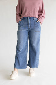 A person wearing blue wide-leg cropped jeans, a dusty pink sweatshirt, and off-white slip-on shoes, standing against a light gray studio background.