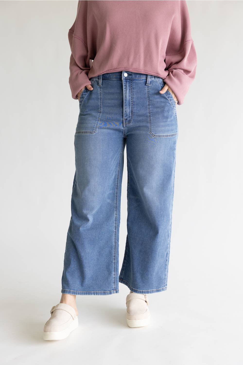 A person wearing blue wide-leg cropped jeans, a dusty pink sweatshirt, and off-white slip-on shoes, standing against a light gray studio background.