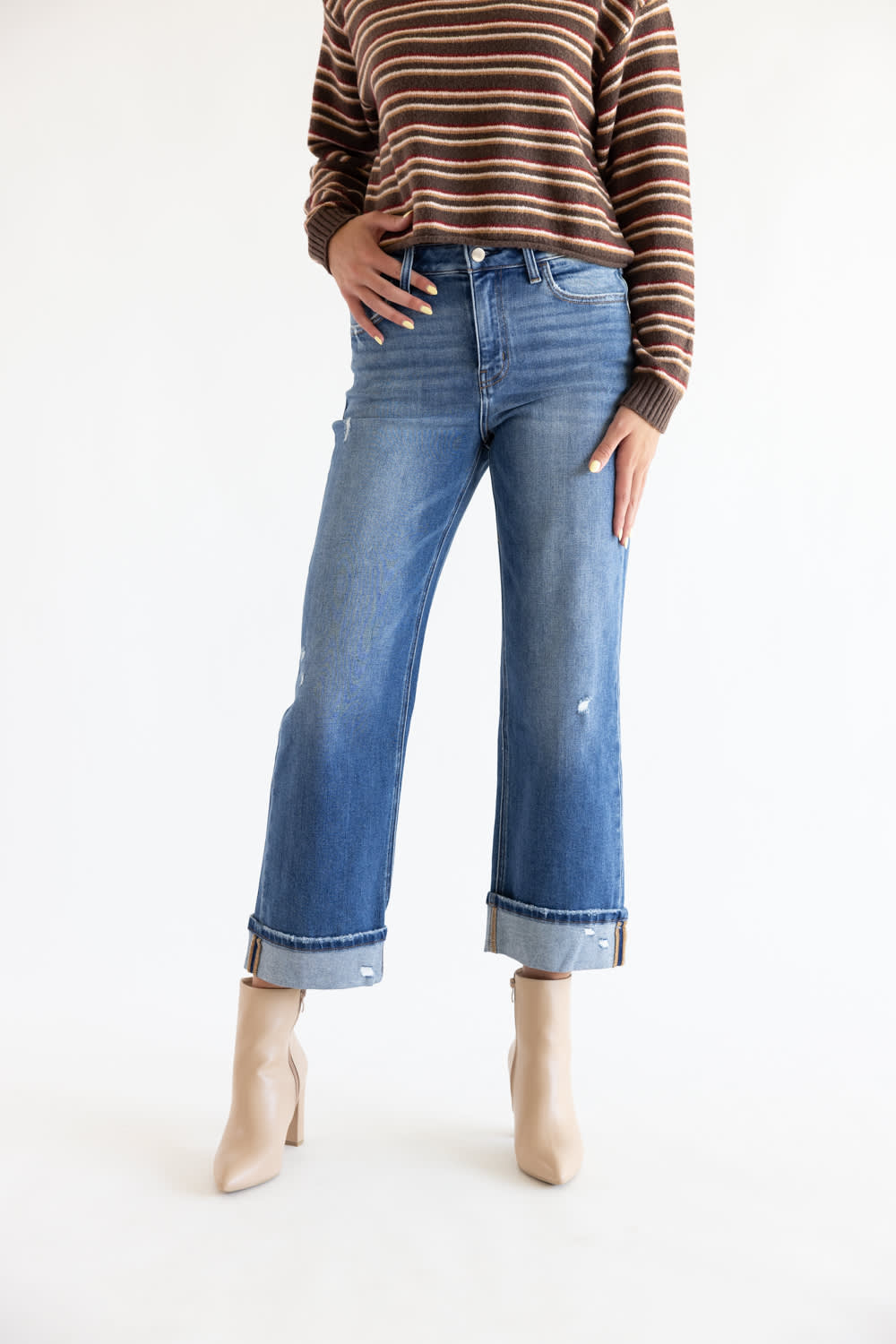 A model wearing blue cropped jeans with rolled cuffs, paired with a brown and beige striped sweater and beige ankle boots, shown against a plain white studio background.