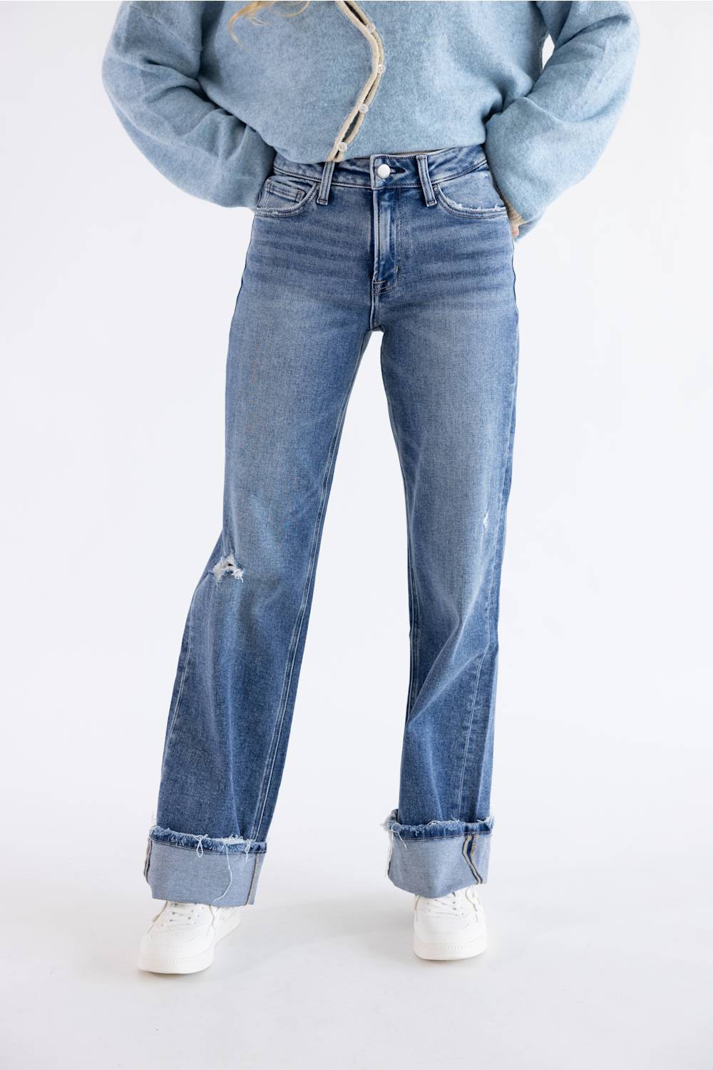 A person wearing light-to-medium wash blue jeans with a distressed knee detail and rolled cuffs, paired with a light blue cropped jacket and white sneakers, against a plain white studio background.