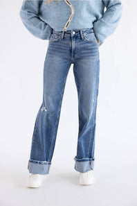 A person wearing light-to-medium wash blue jeans with a distressed knee detail and rolled cuffs, paired with a light blue cropped jacket and white sneakers, against a plain white studio background.