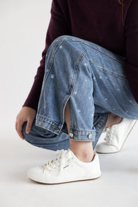 A person crouching in light blue denim jeans with small white dot patterns, paired with white low-top sneakers and a dark burgundy sweater, against a plain white studio background.