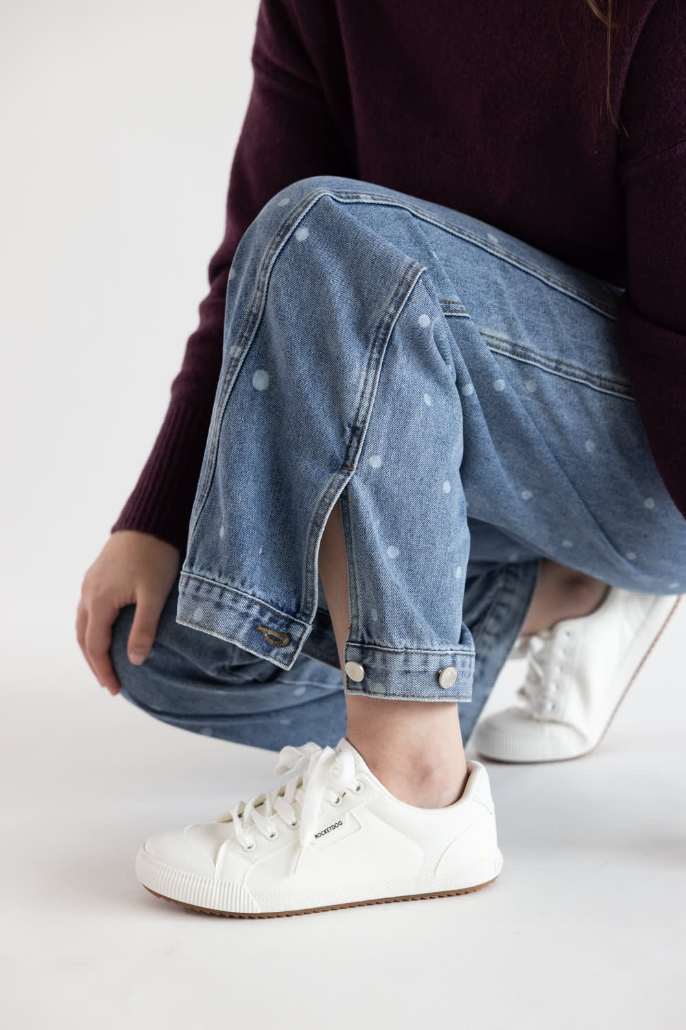 A person crouching in light blue denim jeans with small white dot patterns, paired with white low-top sneakers and a dark burgundy sweater, against a plain white studio background.
