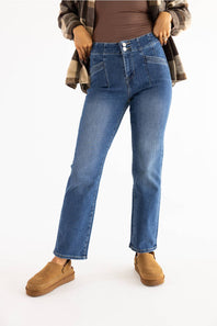 A person wearing medium-wash blue jeans with a brown fitted top, a brown-and-tan plaid flannel shirt, and tan slip-on shoes, photographed against a plain white studio background.