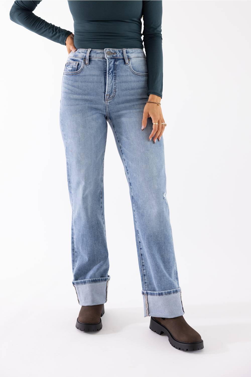 A model wearing light-wash blue denim jeans with rolled cuffs, paired with a dark green long-sleeve top and brown boots, shown from the waist down on a white studio background.