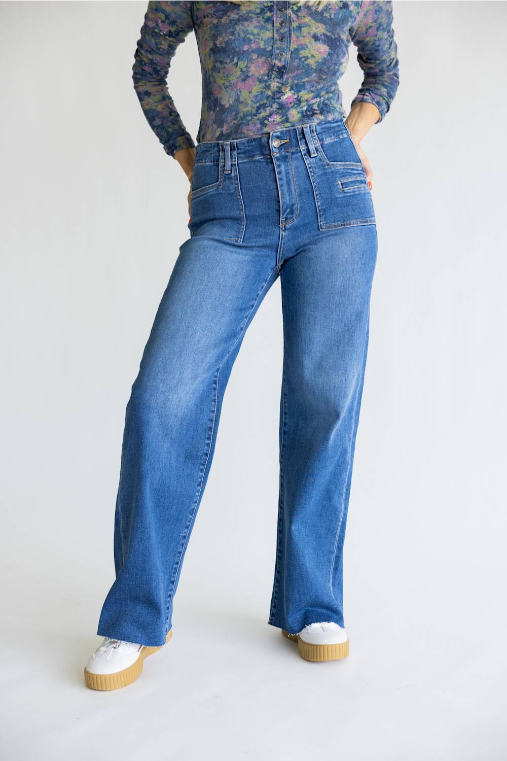 A model wearing a pair of high-waisted, wide-leg blue denim jeans with a floral long-sleeve top and white platform sneakers, standing against a plain white studio background.