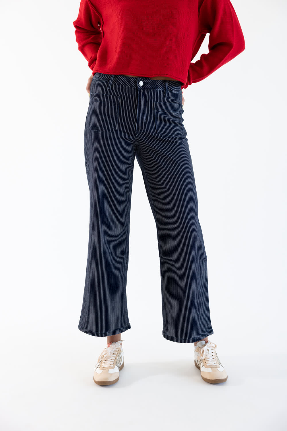 A person wearing dark navy corduroy cropped flare pants, a red long-sleeve top, and beige sneakers, standing against a plain white studio background.