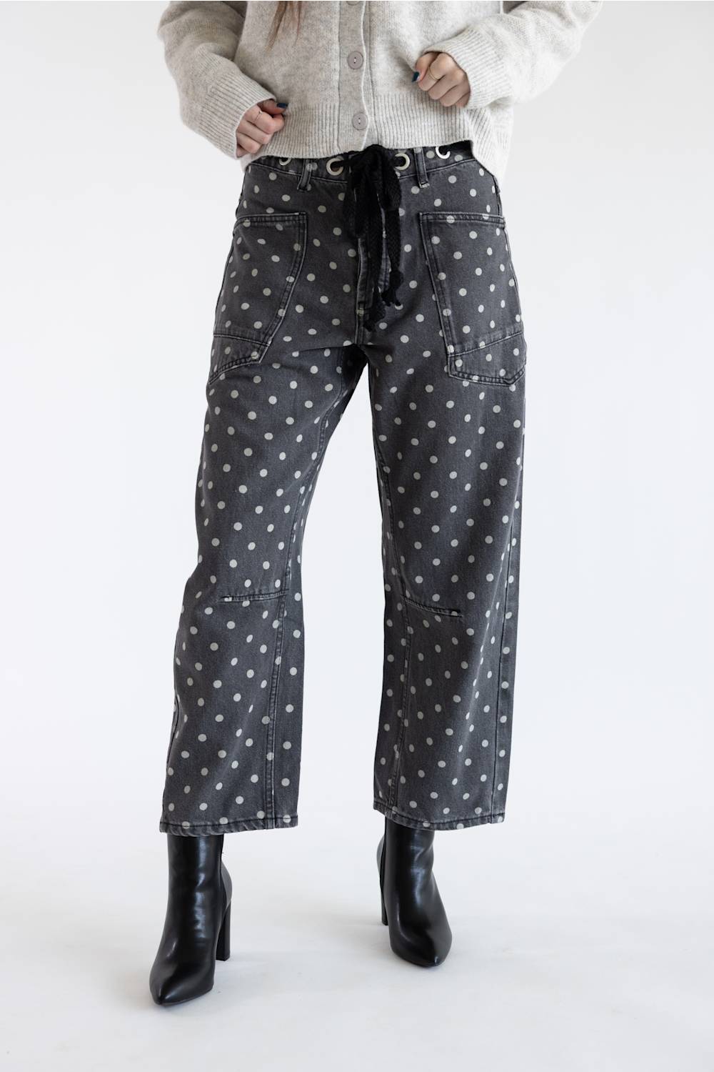 A model posed from the waist down wearing black barrel-leg jeans featuring a white polka dot pattern, black drawstring eyelet waistband, and dual front pockets, styled with black leather heeled boots on a plain white background.